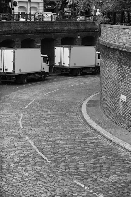 A black-and-white photograph of a narrow, curved cobblestone street leading to a loading area beneath an elevated brick archway. Two large white moving trucks with open rear doors are parked side by s